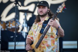 Male bassist wearing a tropical shirt and cap, playing a wooden bass on stage at a live show.