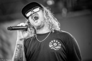 Black-and-white photo of a male singer wearing a cap and sunglasses, shouting into a handheld microphone on stage.