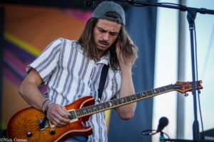 Male guitarist wearing a striped shirt and backwards cap, playing a sunburst electric guitar on stage.