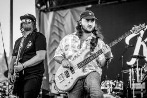 Live concert shot of two male musicians: center-stage bassist with long hair, beard, hat, and a patterned shirt playing a bass; another guitarist in a cap and sunglasses stands to the left.