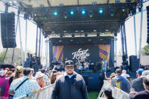 Outdoor festival stage with a live band performing, large speakers and blue stage lights, and a crowd leaning on barriers in front of the stage.