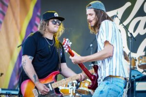Two rock musicians on stage: one wearing a black shirt, cap and sunglasses, playing a red electric guitar while the other in a striped shirt and backwards cap leans in with a guitar on stage.