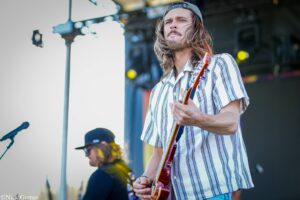 Guitarist performing on a stage, wearing a striped shirt and backwards cap, playing a red electric guitar.