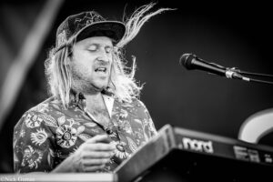 Black-and-white photo of a male musician with long hair and a patterned shirt singing into a microphone while playing a keyboard on stage.