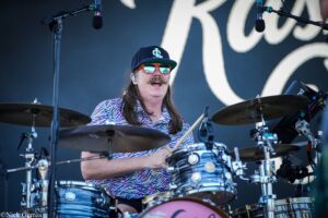 Drummer with long hair, mustache, and orange sunglasses performs on stage behind a blue striped drum kit with microphones nearby.