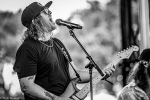 Black-and-white photo of a guitarist on stage, singing into a microphone while playing an electric guitar, wearing a cap and sunglasses on a sunny performance day.
