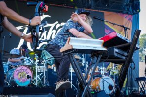 Keyboardist playing a red Yamaha keyboard on an outdoor stage, wearing a patterned shirt and cap with drums visible in the background.
