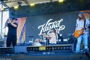 Band performing on outdoor stage with guitarist and vocalist, big screen backdrop and yellow stage lights behind them