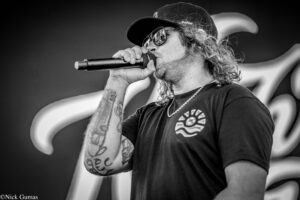 Male singer with long hair and tattoos wearing a cap and sunglasses, singing into a microphone on stage.