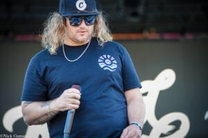Man with long wavy hair, sunglasses, and a navy t‑shirt holding a microphone at a stage or event.