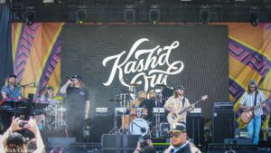 Band performing on a colorful outdoor stage with a large screen behind them displaying a white script logo and multiple musicians playing instruments.