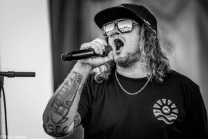 Male singer wearing a cap and mirrored sunglasses, gripping a mic and singing on stage; tattooed forearm visible.