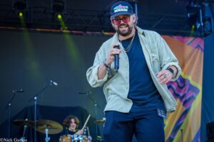 Male singer on stage singing into a handheld microphone, wearing a cap, sunglasses, and a beige button-up shirt over a dark tee.