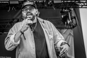 Male singer with glasses and a beard, wearing a striped shirt, performing into a handheld microphone on stage.
