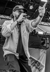 Male vocalist on stage, wearing a cap and glasses, holding a microphone and pointing upward with his other hand, stage rigging in the background.