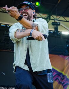 Male performer on stage pointing at the crowd, holding a microphone, wearing a cap, glasses, and a light green striped shirt.