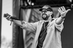 Male singer performing on stage, microphone in hand, wearing a cap and glasses, smiling with arms outstretched