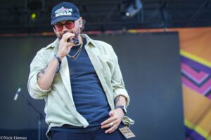 Male singer on stage wearing a navy cap with a fish logo, sunglasses, and a beige shirt over a dark T-shirt, holding a microphone.