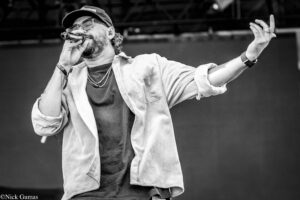 Male singer performing on stage, mic near mouth, wearing cap, glasses, and necklace in a black-and-white photo, gesturing with his right hand raised.