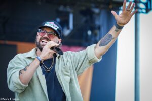 Male singer wearing a cap and sunglasses sings into a microphone with his right hand raised on stage.