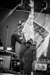 Male vocalist on stage mid-jump, microphone in hand, one arm raised, wearing a cap and glasses with equipment in the background (black and white).