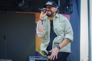 Male singer on stage, wearing a cap and glasses, smiling while holding a microphone close to his mouth during a live performance.