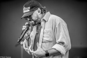 Male singer with a cap and sunglasses singing into a handheld microphone on stage, in a striped shirt.