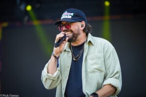 Male singer on stage, holding a microphone, wearing a dark cap, pink sunglasses, and a beige corduroy shirt.