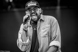 Black-and-white photo of a man singing into a microphone on stage, wearing a cap, glasses, and a striped shirt in a lively performance moment.