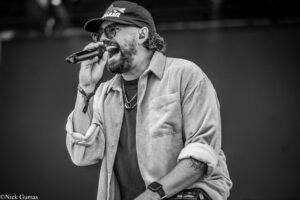 Black-and-white photo of a male singer performing on stage, wearing a cap, sunglasses, and a striped shirt while singing into a handheld microphone.