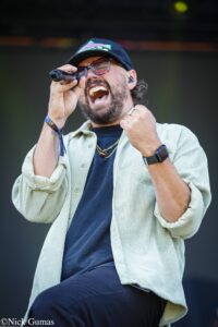 Male singer wearing a cap and glasses, passionately singing into a handheld microphone on stage.