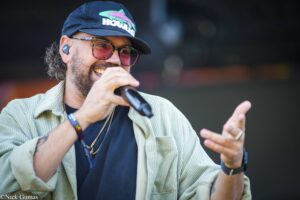 Male singer performing into a microphone, wearing a cap, sunglasses, and a light corduroy jacket on stage.