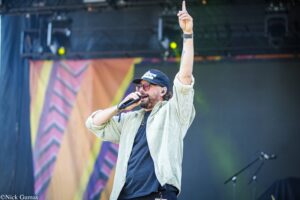 Male singer on stage raises his right index finger while singing into a microphone, wearing a cap and glasses.