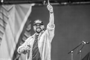 Male performer on a stage raising his right index finger in the air, holding a microphone in his left hand, wearing a cap, glasses, and a striped jacket.