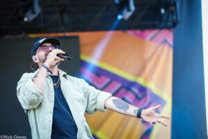 Male singer wearing a cap and sunglasses sings into a microphone on a colorful stage backdrop