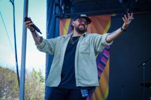 Male singer on an outdoor stage, arms wide open, holding a microphone in his left hand, wearing a cap and glasses.