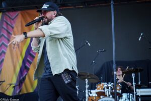 Male singer on stage, wearing a cap and glasses, singing into a handheld microphone with a drummer visible in the background.