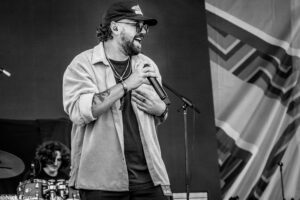 Male singer with a beard and cap laughing into a handheld microphone on stage, with a drummer in the background (black-and-white).