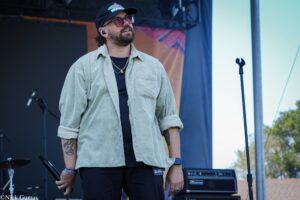 Male performer on an outdoor stage, wearing a light green shirt, black shirt, cap, sunglasses, in-ear monitor, holding a microphone with stage gear in the background.