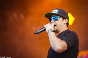 Male singer wearing a black cap and blue mirrored sunglasses, singing into a microphone on stage with an orange background behind him.