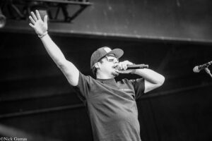 Male singer on stage, wearing a cap and sunglasses, raising one arm while singing into a microphone.