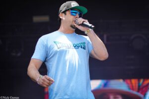 Male singer performing on stage, holding a microphone, wearing a cap and blue sunglasses, in a light blue T-shirt.