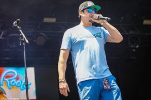 Male performer singing into a microphone on stage, wearing a gray cap, blue sunglasses, and a light blue Cali Roots shirt.