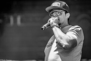 Male performer singing into a microphone on stage, wearing a cap and reflective sunglasses, monochrome photo