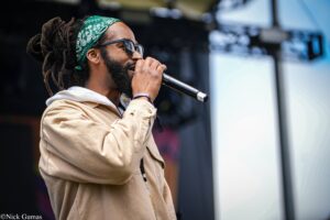 Male singer with dreadlocks and a green bandana singing into a microphone on stage, wearing sunglasses and a beige jacket.