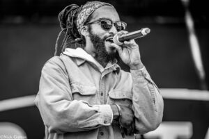 Black-and-white photo of a male singer with dreadlocks, a bandana, and sunglasses singing into a handheld microphone.