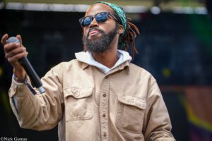 Male performer with sunglasses and a green bandana sings into a microphone on stage.