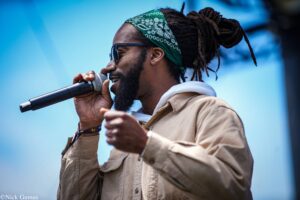 Male singer wearing a green bandana and sunglasses, singing into a handheld microphone on a blue backdrop stage.