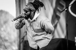 Black-and-white photo of a bearded man singing into a microphone on stage, wearing a bandana and sunglasses.