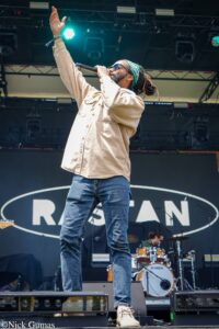 Male singer on stage raising one arm, holding a mic, wearing a beige shirt and green bandana, performing with drums in the background.
